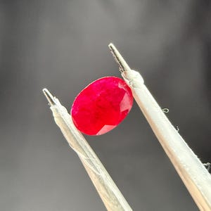 Puede incluir: Un rubí ovalado, facetado, de color rojo intenso, sostenido por unas pinzas plateadas sobre un fondo oscuro. La gema muestra un color rojo saturado y una excelente claridad. Las pinzas están inclinadas, mostrando la gema.