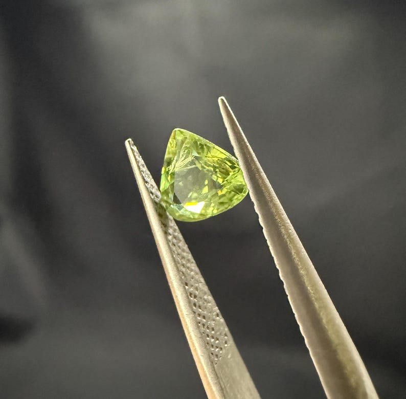 Pode incluir: Uma gema verde com forma triangular &eacute; segurada por uma pin&ccedil;a.