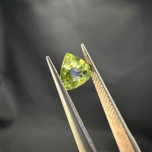 Pode incluir: Uma gema verde com formato triangular &eacute; segurada por uma pin&ccedil;a. A gema tem um leve brilho.