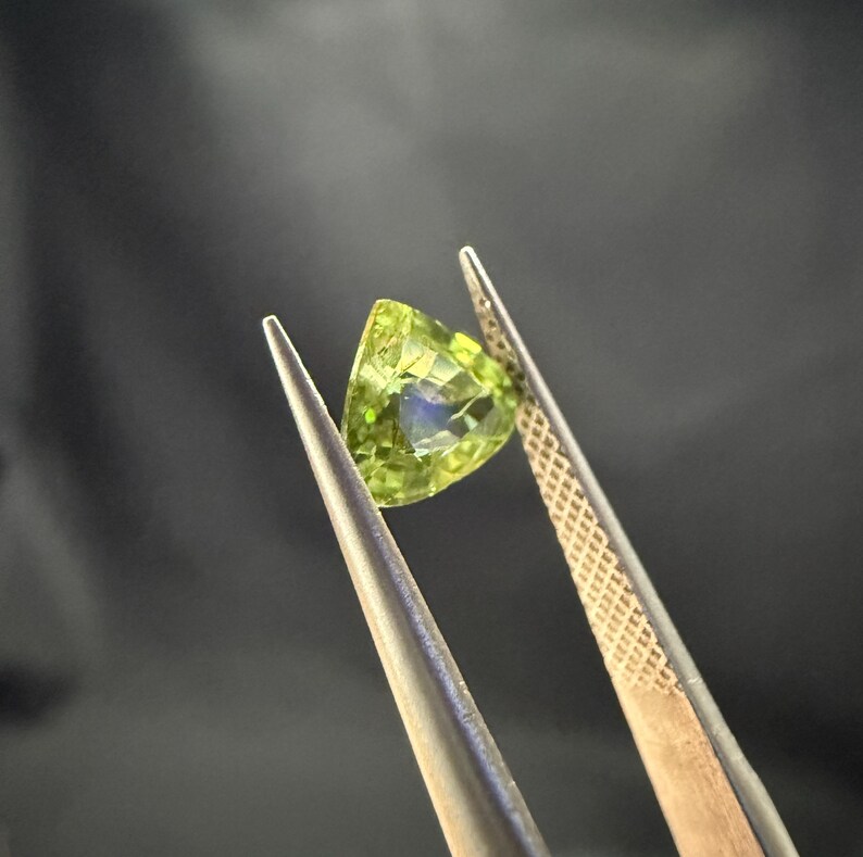 Pode incluir: Uma gema verde com formato triangular &eacute; segurada por uma pin&ccedil;a. A gema tem um leve brilho.