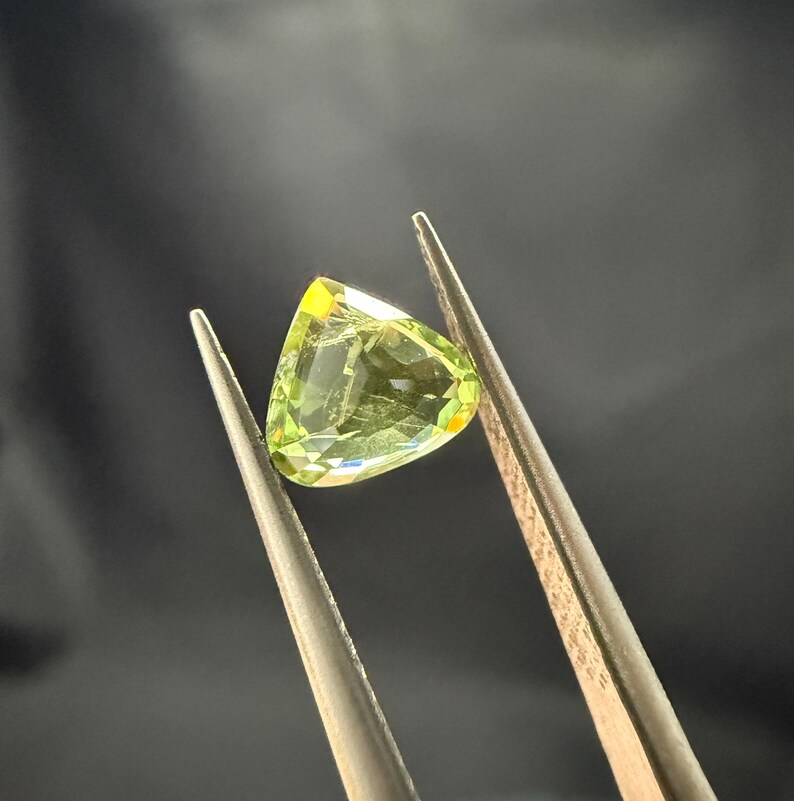 Pode incluir: Uma gema verde com formato triangular, segurada por uma pin&ccedil;a.