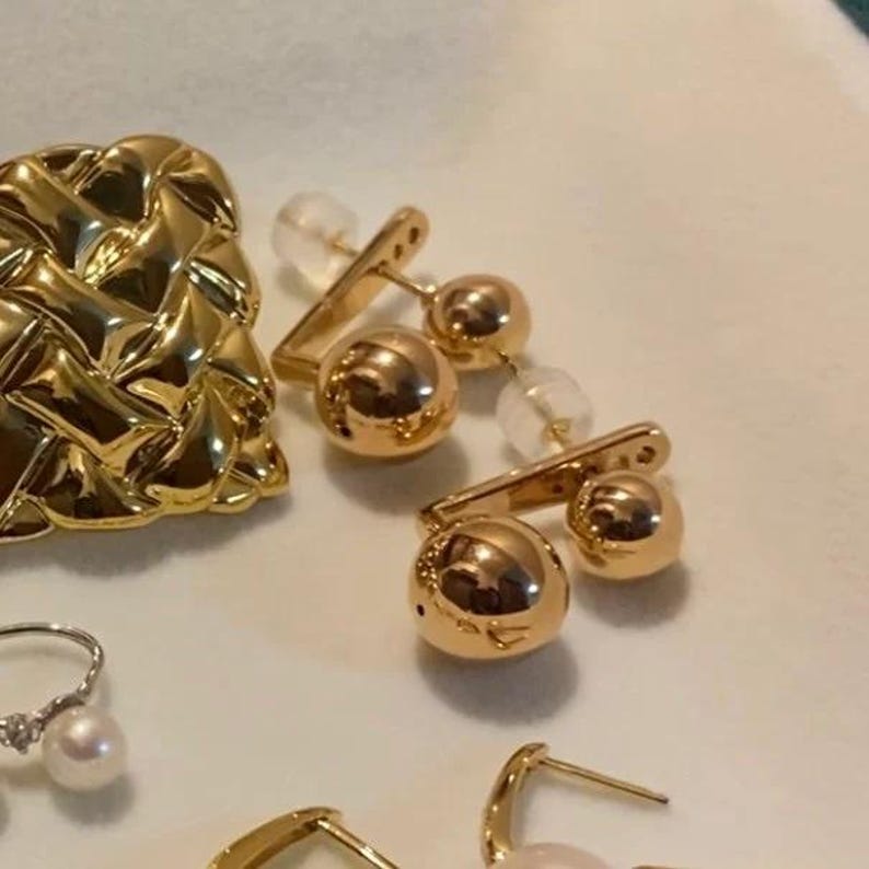 Puede incluir: Un par de pendientes de tono dorado con dos bolas redondas en cada pendiente. Los pendientes est&aacute;n hechos de metal y tienen un acabado pulido.