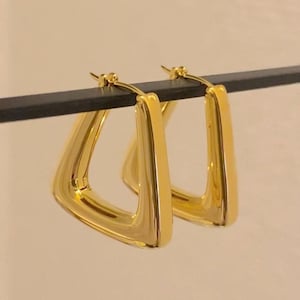 Puede incluir: Un par de pendientes de aro de forma triangular de color dorado. Los pendientes están hechos de metal grueso y tienen un acabado liso y pulido.