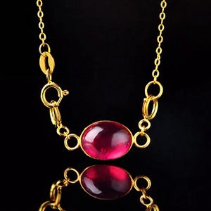 以下が含まれることがあります： 大きな楕円形のルビーレッドの宝石ペンダントが付いた、ゴールドカラーのネックレス。 ネックレスは繊細なチェーンとクラスプクロージャーが特徴です。 宝石はゴールドベゼルにセットされており、ネックレスは黒い背景に表示されています。