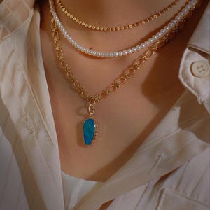 May include: A gold chain necklace with a blue opal pendant. The necklace is layered with a pearl necklace and a thin gold chain.