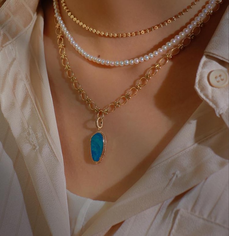 May include: A gold chain necklace with a blue opal pendant. The necklace is layered with a pearl necklace and a thin gold chain.