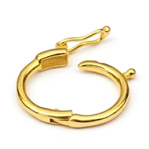 May include: A gold-coloured, circular jewellery clasp, open and ready to be closed. The clasp has a spring-loaded lever and a small, rounded knob on the opposite side. The metal has a shiny finish, reflecting light. The clasp is designed for jewellery making.