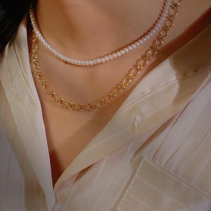 May include: A layered necklace with a delicate pearl strand and a gold chain with a repeating floral design.