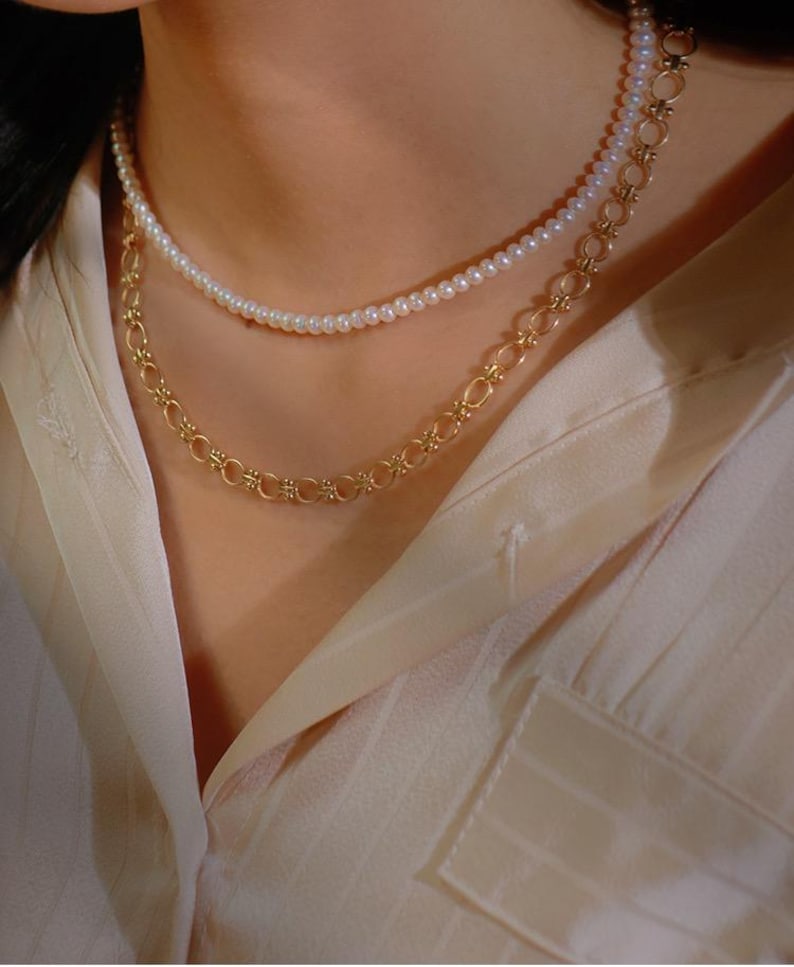 May include: A layered necklace with a delicate pearl strand and a gold chain with a repeating floral design.