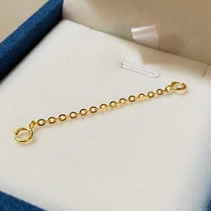 May include: A gold-colored necklace extender with a delicate chain of small, round links. The extender has a spring ring clasp on each end, designed to lengthen necklaces or bracelets. The piece is presented in a blue jewelry box with a white interior.