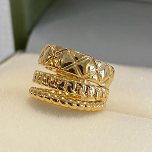 May include: Three gold rings stacked together. The top ring has a quilted pattern, the middle ring has a chain-like design, and the bottom ring has a rope-like texture. The rings are displayed on a white surface.