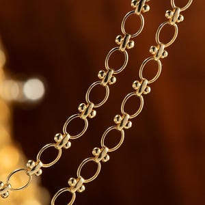 May include: A close-up of a gold chain necklace with oval links and small round beads. The chain is layered on a brown background.