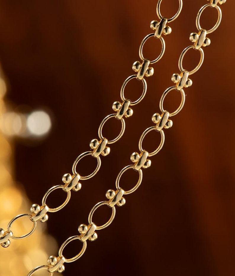 May include: A close-up of a gold chain necklace with oval links and small round beads. The chain is layered on a brown background.