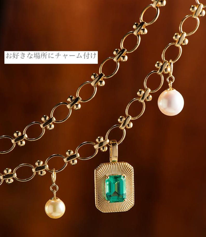 May include: A gold chain necklace with three charms: a green emerald gemstone pendant, a white pearl, and a smaller white pearl. The chain is made of interlocking circles with small beads between each circle.