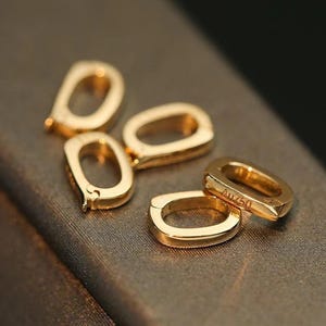 Puede incluir: Cuatro anillos de salto de metal de color dorado, uno de los cuales tiene la inscripción "Au750" en el lateral. Los anillos están sobre una superficie marrón.