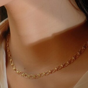May include: A gold chain necklace with a delicate, intricate design. The chain is made up of small, linked ovals with a textured surface.