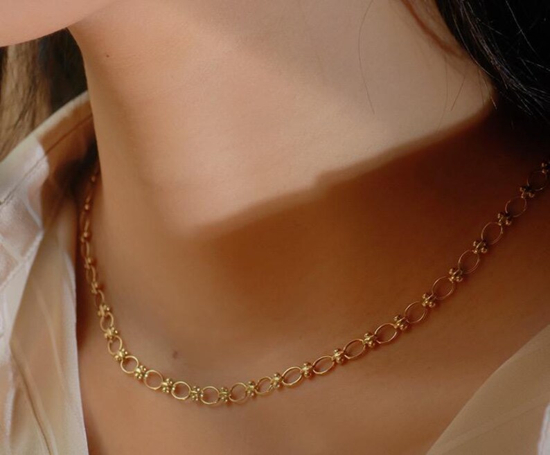 May include: A gold chain necklace with a delicate, intricate design. The chain is made up of small, linked ovals with a textured surface.
