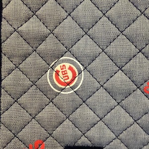 Könnte beinhalten: Nahaufnahme eines gesteppten Stoffes mit Rautenmuster in Grau- und Marineblautönen. Der Stoff ist mit roten "Cubs"-Logos und -Texten verziert, die sich in weißen Kreisen befinden. Die Nähte sind schwarz.