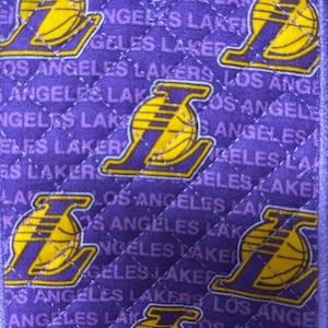 May include: Purple quilted fabric featuring the Los Angeles Lakers logo in yellow and purple. The words "LOS ANGELES LAKERS" are printed repeatedly across the fabric. The design has a diagonal quilted pattern.