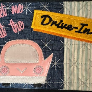 Puede incluir: Un coche rosa con un parabrisas blanco y un letrero amarillo que dice "Drive-In". El fondo es una tela estampada azul y blanca. El texto "Meet me at the" está bordado en rosa en la tela.