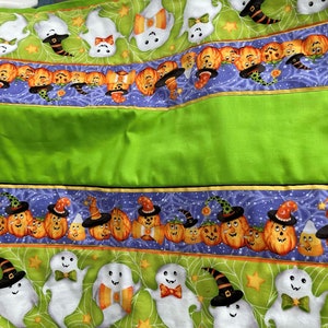 May include: A green fabric with two rows of Halloween-themed fabric trim. The top row features white ghosts with black hats and bowties. The bottom row features orange pumpkins with black witch hats and candy corn.