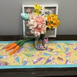 Może przedstawiać: Żółty bieżnik na stół w tematyce wielkanocnej z powtarzającym się wzorem gnomów w czapkach w kształcie rożków lodowych. Turkusowa obwódka. Szklany słoik zawiera kwiaty i kolorowe cukierki. Obecne są również sztuczne marchewki i pisanka.