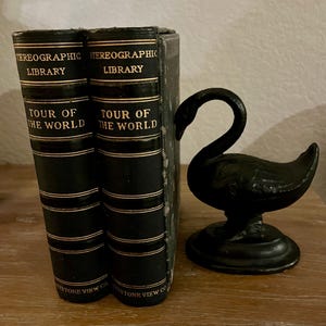 May include: Two vintage black books titled "Tour of the World" from the Stereographic Library, with gold lettering and accents. A black swan figurine sits beside the books. The books are from Keystone View Co.