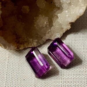May include: Two faceted, rectangular amethyst gemstones with a deep purple hue. The gemstones are displayed on a textured, off-white surface, with a portion of a rough, light-colored mineral formation in the background.