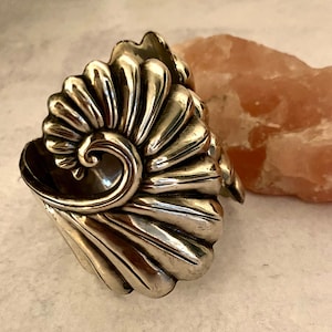 May include: A silver cuff bracelet with a detailed design of a stylized flower or shell. The bracelet is made of a smooth, polished metal and has a slightly curved shape.