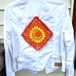 May include: White denim jacket with a crocheted sun design on the back. The sun is a vibrant mix of orange, yellow, and red, set against the clean white fabric. The jacket has classic denim details.