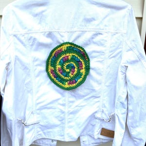 May include: White denim jacket with a large, colorful, crocheted spiral design on the back. The spiral features green, yellow, pink, purple, and blue yarn. The jacket has silver hardware and a small tag.