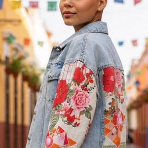 May include: Light blue denim jacket with floral patterned sleeves. The sleeves feature a vibrant design of red roses, pink flowers, and geometric shapes on a cream background. The jacket has a classic button-up design.
