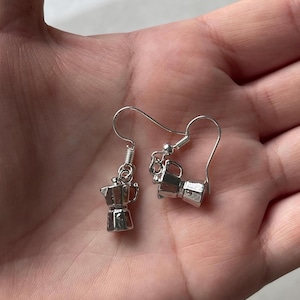 May include: A pair of silver-colored earrings featuring miniature coffee pots. The earrings have a hook design for pierced ears. The coffee pot charms are detailed with a classic Italian stovetop espresso maker design.