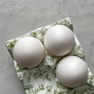 Peut inclure: Un coquetier en c&eacute;ramique blanche orn&eacute; d'un motif botanique et de papillons verts, contenant trois &oelig;ufs blancs lisses et mats. Le plateau pr&eacute;sente des empreintes moul&eacute;es pour les &oelig;ufs et une forme rectangulaire aux bords incurv&eacute;s.