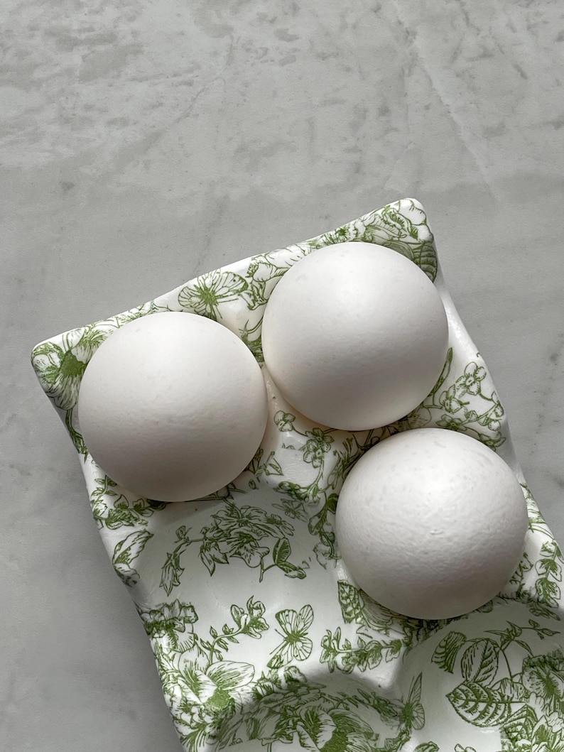 Peut inclure: Un coquetier en c&eacute;ramique blanche orn&eacute; d'un motif botanique et de papillons verts, contenant trois &oelig;ufs blancs lisses et mats. Le plateau pr&eacute;sente des empreintes moul&eacute;es pour les &oelig;ufs et une forme rectangulaire aux bords incurv&eacute;s.