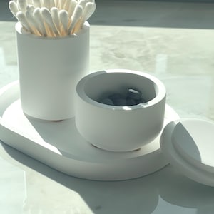 May include: A white bathroom vanity set includes a cylindrical container filled with cotton swabs, a small round container with gray stones, and a matching oval tray. The set is made of a matte white material.