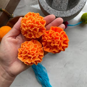 May include: Three bright orange marigold flower-shaped objects with intricate, ruffled petal textures. Each flower features a dense, layered design with a rounded, spherical shape, showcasing a vibrant, solid orange color throughout the entire surface.