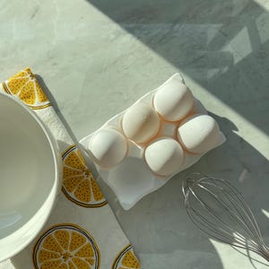 Peut inclure: Un plateau &agrave; &oelig;ufs en c&eacute;ramique blanche contenant cinq gros &oelig;ufs blancs, plac&eacute; &agrave; c&ocirc;t&eacute; d'un bol m&eacute;langeur blanc, d'un fouet en m&eacute;tal et d'un torchon blanc orn&eacute; d'un motif r&eacute;p&eacute;titif de tranches de citron jaunes et noires.