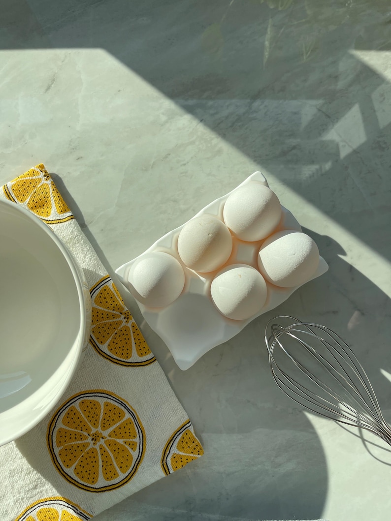 Peut inclure: Un plateau &agrave; &oelig;ufs en c&eacute;ramique blanche contenant cinq gros &oelig;ufs blancs, plac&eacute; &agrave; c&ocirc;t&eacute; d'un bol m&eacute;langeur blanc, d'un fouet en m&eacute;tal et d'un torchon blanc orn&eacute; d'un motif r&eacute;p&eacute;titif de tranches de citron jaunes et noires.