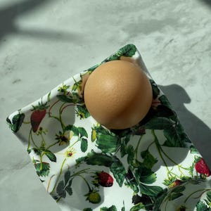 Peut inclure: Un coquetier en c&eacute;ramique blanche orn&eacute; d'un imprim&eacute; botanique d&eacute;taill&eacute; repr&eacute;sentant des feuilles de fraisier vertes, des fleurs blanches et des fraises rouges. Un &oelig;uf brun repose dans l'un des creux circulaires du plateau rectangulaire.