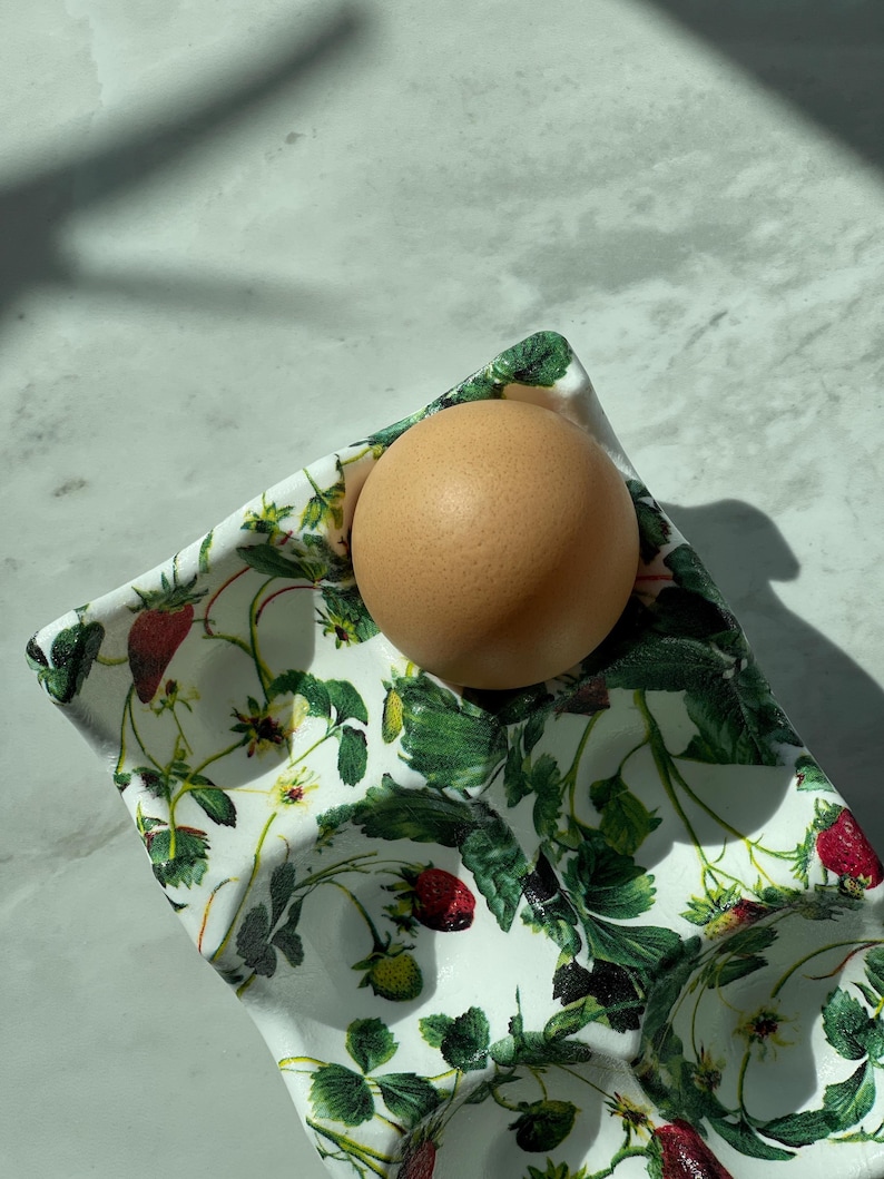 Peut inclure: Un coquetier en c&eacute;ramique blanche orn&eacute; d'un imprim&eacute; botanique d&eacute;taill&eacute; repr&eacute;sentant des feuilles de fraisier vertes, des fleurs blanches et des fraises rouges. Un &oelig;uf brun repose dans l'un des creux circulaires du plateau rectangulaire.