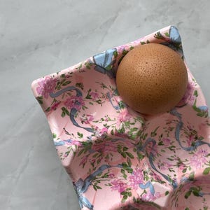 Peut inclure: Une bo&icirc;te &agrave; &oelig;ufs rose orn&eacute;e d'un motif r&eacute;p&eacute;titif de rubans bleu clair et de grappes de petites fleurs roses avec des feuilles vertes. Un &oelig;uf brun est plac&eacute; dans l'un des compartiments moul&eacute;s de la bo&icirc;te, qui pr&eacute;sente une finition textur&eacute;e.