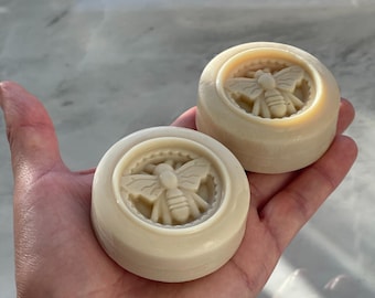 May include: Two round, off-white soaps with a bee design. The bee is in the center of the soap, surrounded by a circular border. The soaps are held in a hand, and the background is a light, marble-like surface.