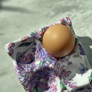 Peut inclure: Un coquetier en c&eacute;ramique blanche orn&eacute; d'un motif floral color&eacute; compos&eacute; de fleurs de lilas violettes et de petits papillons bleus. Un &oelig;uf de poule brun est nich&eacute; dans l'un des compartiments circulaires moul&eacute;s du plateau.