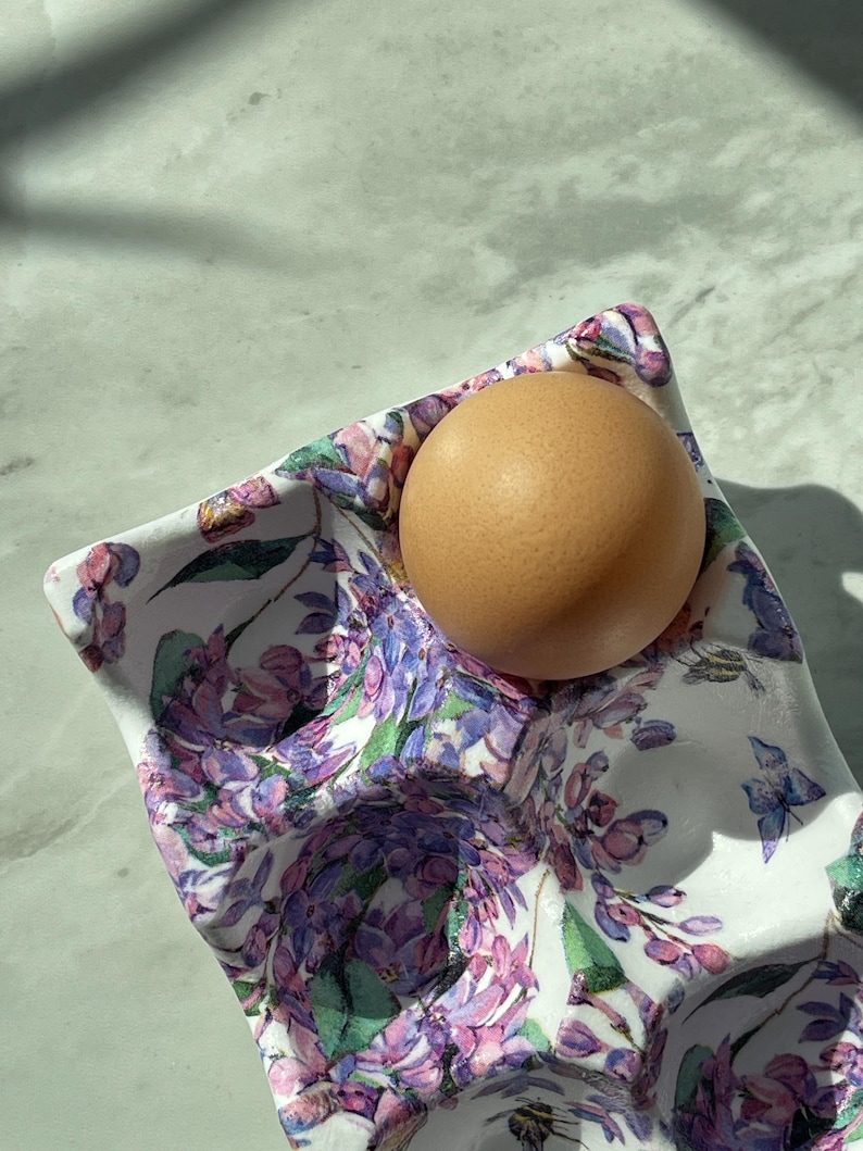 Peut inclure: Un coquetier en c&eacute;ramique blanche orn&eacute; d'un motif floral color&eacute; compos&eacute; de fleurs de lilas violettes et de petits papillons bleus. Un &oelig;uf de poule brun est nich&eacute; dans l'un des compartiments circulaires moul&eacute;s du plateau.