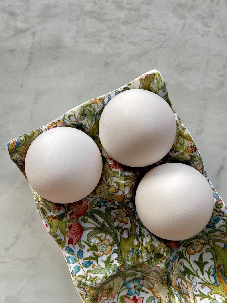 Peut inclure: Trois &oelig;ufs blancs mats et lisses sont nich&eacute;s dans un support en c&eacute;ramique d&eacute;coratif. Le support pr&eacute;sente un motif botanique d&eacute;taill&eacute; et color&eacute; avec des feuilles vertes, des fleurs orange et des touches bleues sur fond blanc.