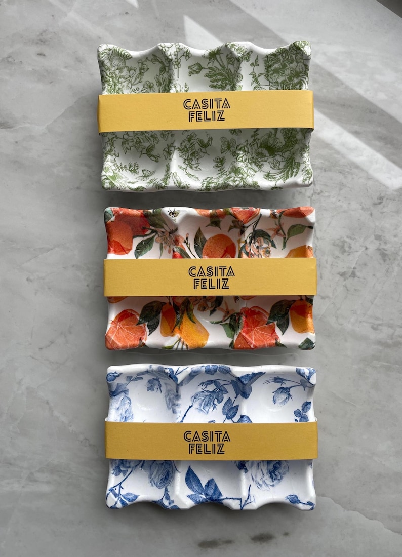 May include: Three ceramic serving trays with floral patterns and the text "CASITA FELIZ" on each. The trays have different designs: green and white floral, orange and yellow citrus, and blue and white roses. Each tray is wrapped with a yellow band.