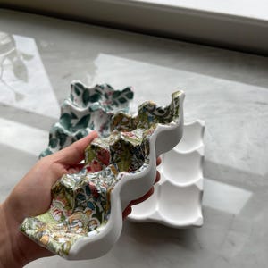 Peut inclure: Une bo&icirc;te &agrave; &oelig;ufs en c&eacute;ramique avec un ext&eacute;rieur blanc et un motif floral et aviaire color&eacute; sur la surface sup&eacute;rieure. Le plateau pr&eacute;sente six empreintes moul&eacute;es pour contenir des &oelig;ufs. Une seconde bo&icirc;te &agrave; &oelig;ufs blanche est visible.