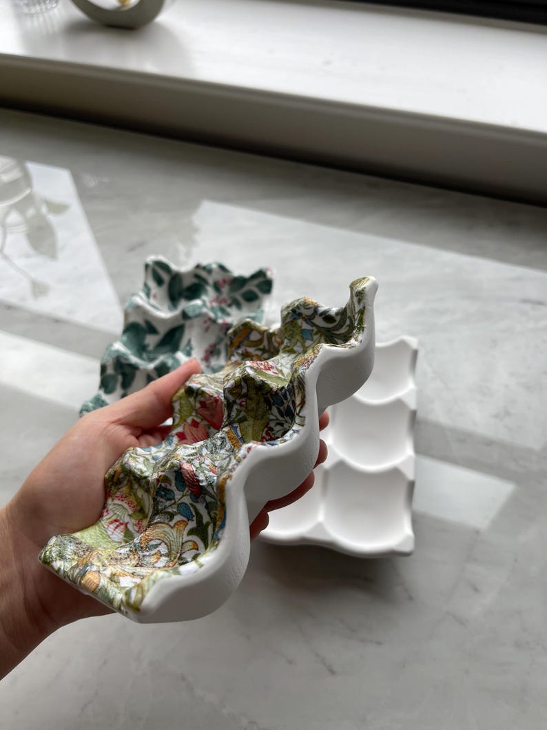 Peut inclure: Une bo&icirc;te &agrave; &oelig;ufs en c&eacute;ramique avec un ext&eacute;rieur blanc et un motif floral et aviaire color&eacute; sur la surface sup&eacute;rieure. Le plateau pr&eacute;sente six empreintes moul&eacute;es pour contenir des &oelig;ufs. Une seconde bo&icirc;te &agrave; &oelig;ufs blanche est visible.