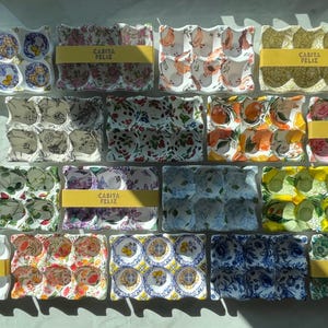 May include: A collection of decorative egg cartons with various floral and patterned designs. Each carton has a yellow band with the text "CASITA FELIZ". The cartons are in shades of white, blue, pink, green, and orange.